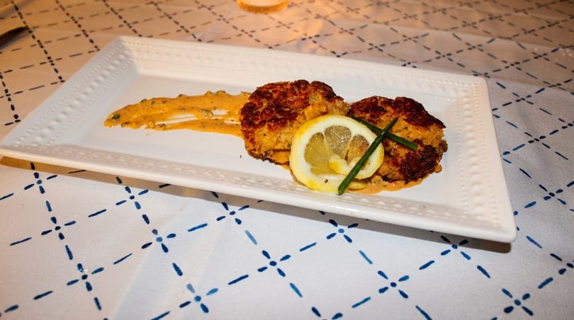 Two golden-brown crab cakes with sauce, lemon, and chives on a white rectangular plate.