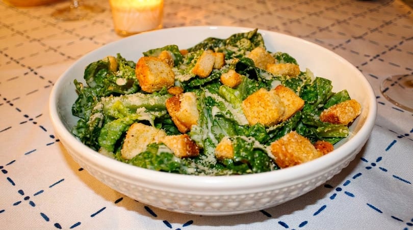 Caesar salad with croutons and parmesan cheese in a white bowl on a patterned tablecloth.