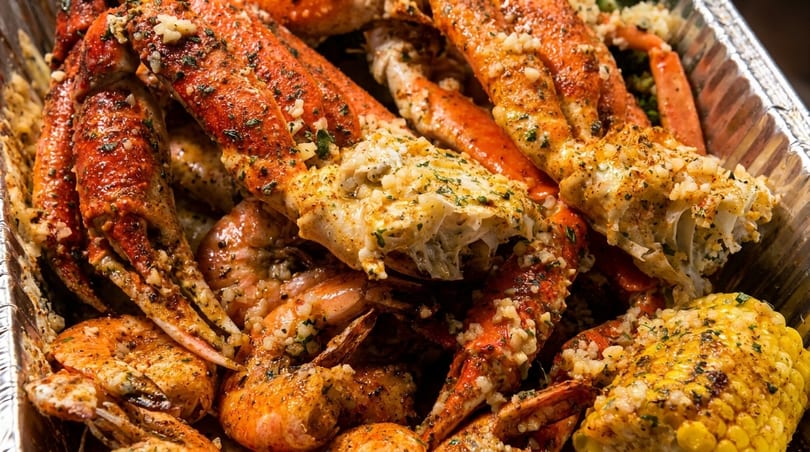 Close-up of a seafood boil with crab legs, shrimp, and corn on the cob in a foil pan.